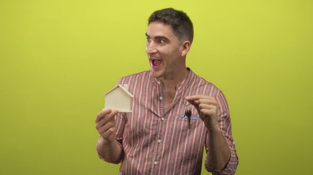 Man holding a wooden house model and metal keys, mouth open in surprise, wearing striped shirt in a lime green studio; home buying excitement.