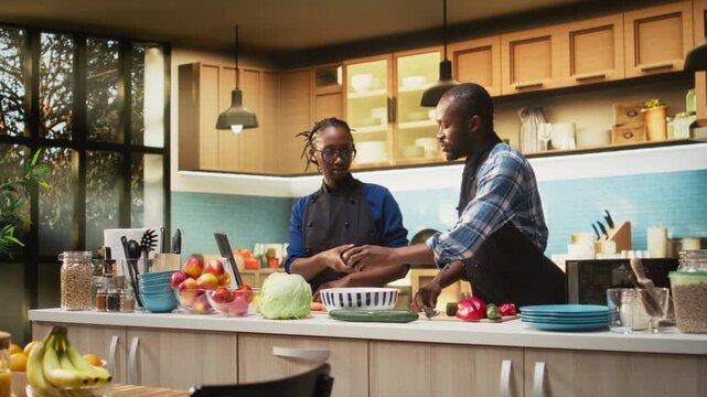 African american boyfriend cutting an avocado to add inside the salad, his girlfriend chopping other vegetables and mixing ingredients in a bowl. Partners preparing vegetarian lunch. Camera A.