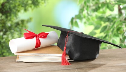 Graduation cap, diploma and notebooks on wooden table against blurred background, closeup