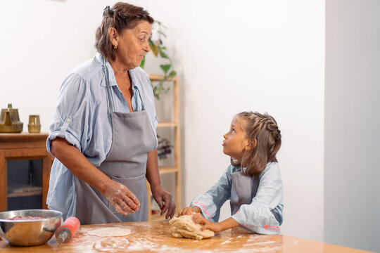Girl and grandmother in aprons working together kneading dough on floured kitchen table enjoying family time, baking and learning at home - Powered by Adobe