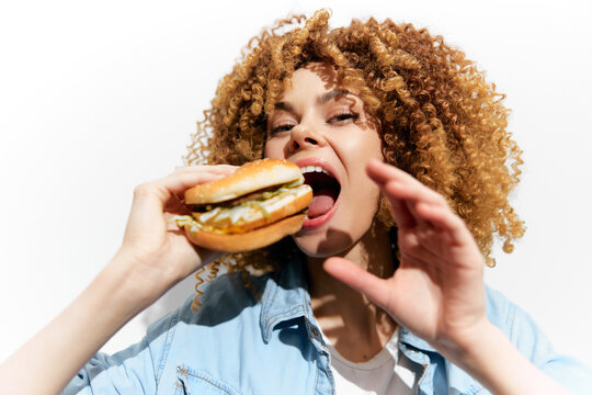 Young woman enjoying a tasty burger with excitement, showcasing her curly hair and casual outfit against a bright white background Perfect for food and lifestyle themes
