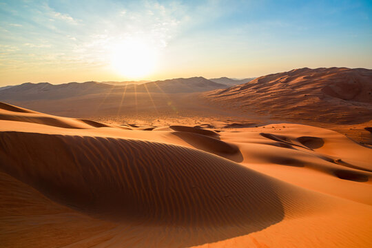 Rub' al-Khali (Empty Quarter) desert in Thumrayt, Dhofar, Oman, presenting sprawling orange sand dunes and a small settlement at the foot of distant hills under a clear morning sky