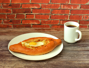 Khachapuri, traditional Georgian pie with egg and cheese in a plate on a wooden table, next to a cup of hot tea.