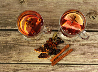 Two traditional glasses of hot red wine with citrus slices and various spices on a wooden table.