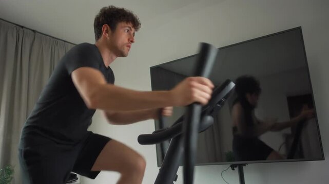 A focused man in black activewear exercising on an elliptical machine in a modern home gym with a large TV screen.