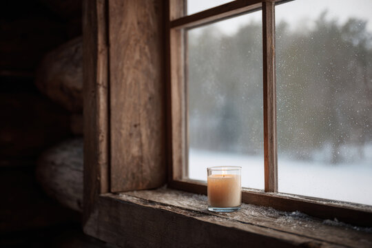 warm candle glowing by rustic wooden window in cozy log cabin during christmas night with gently falling snow - Powered by Adobe