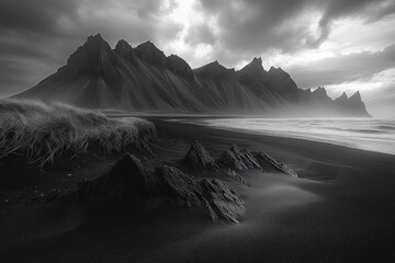 Brooding monochrome coastal landscape with jagged mountain peaks, black sand beach, windswept grass and misty surf under a stormy sky