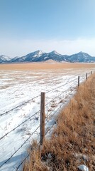 A barbed wire fence runs along a rural landscape covered in patches of snow and dry grass, with snow-capped mountains visible on the horizon.
