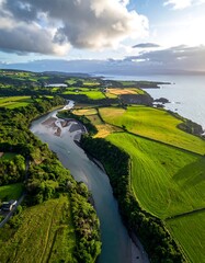 Aerial view of a river winding through vibrant green fields toward a sunlit coast under a cloudy sky