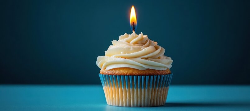 vanilla cupcake in blue liner with swirled buttercream and single lit candle on teal background conveying a quiet celebratory mood - Powered by Adobe