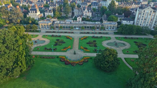 Drone shot descending toward Parc du Thabor in Rennes, revealing French-style gardens, paths, fountains, surrounding buildings and warm sunrise light.