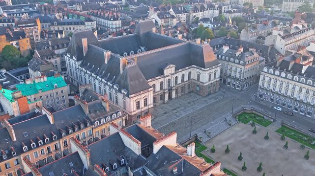 Diagonal drone approach toward the Parlement de Bretagne at sunrise, with a tilt-down revealing the square, rooftops, trees and surrounding streets in Rennes - Brittany