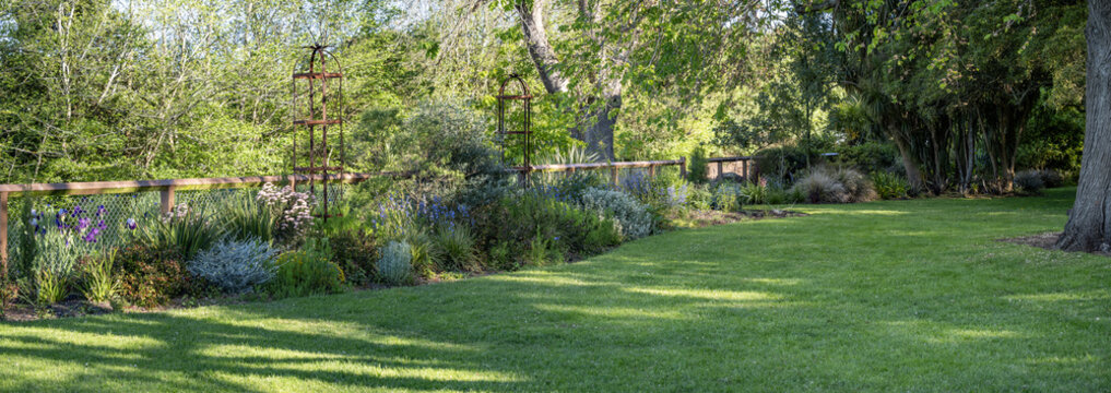 Panoramic background texture of a lush garden with a neat green lawn and a well-defined planted border along the fence line, featuring layered shrubs, flowers, and leafy trees. Landscaping design.