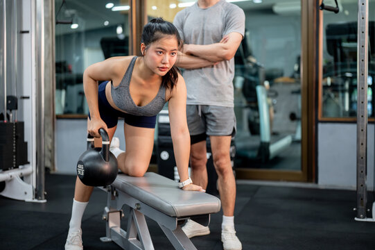 Pretty asian woman client holding kettlebell exercising on bench and fitness trainer coaching in gym