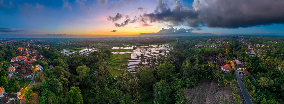 Aerial drone view of water-filled rice paddies reflecting the sunset sky, surrounded by a traditional Balinese village and coconut trees