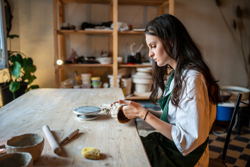 Molding handmade ceramics. Table in pottery studio with female artist shaping wet clay for potter pot or vase. Ceramist master or craftswoman shaping kitchenware in creative workplace