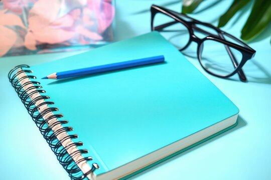 turquoise spiral notebook with blue pencil and black eyeglasses on a pastel blue desk beside a floral container and green leaves, calm inviting study scene