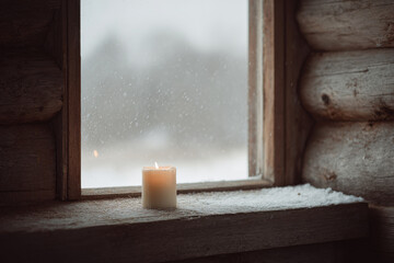 warm candle glowing by rustic wooden window in cozy log cabin during christmas night with gently falling snow