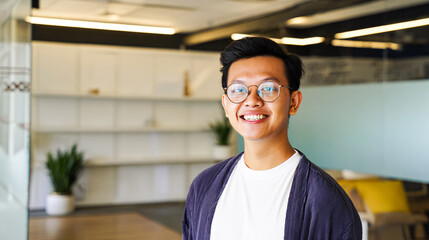 happy young Asian man in glasses standing in a modern office. Confident creative professional or startup employee looking at camera with a smile.