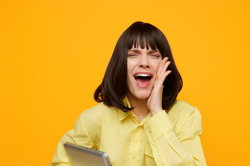 Bright portrait of a joyful young woman in a yellow shirt against a vivid orange backdrop, laughing with mouth open and a hand near her face, holding a notebook for lively lifestyle and education