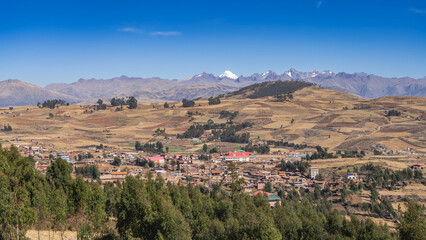 Obraz premium Rural houses, agricultural fields, and green trees are visible on the high-altitude plateau. A mountain range with snow-capped peaks against a clear blue sky. Peru. The Andes Mountains.