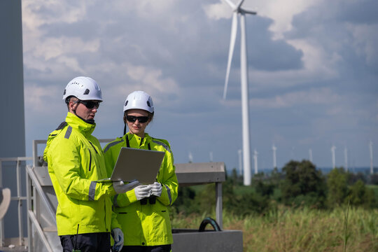 Workers at a wind farm analyze data on a laptop while wearing safety gear under a cloudy sky - Powered by Adobe