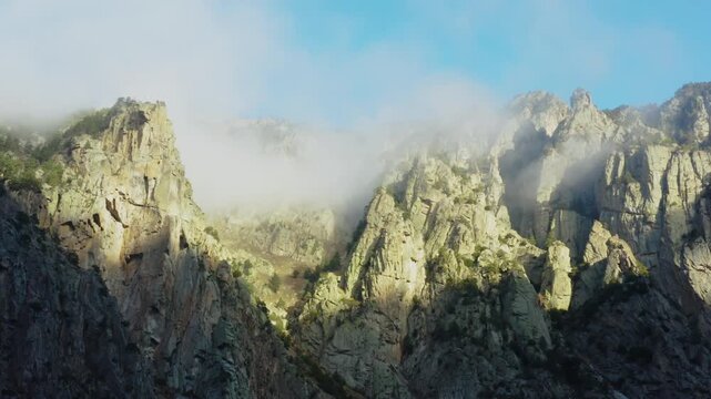 A stunning view of the rugged, rocky peaks of the North Caucasus mountains in Russia. Sunlight illuminates the dramatic landscape as wispy clouds gracefully drift across the sky and through the valley