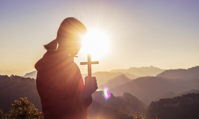 Girl praying and holding Christian cross for worshipping God on mountain at sunrise background. Christian, Christianity, Religion copy space background. Easter Sunday concept: