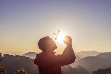 Girl praying and holding Christian cross for worshipping God on mountain at sunrise background. Christian, Christianity, Religion copy space background. Easter Sunday concept: