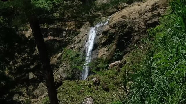 A steady 50-meter waterfall flows down the rocky cliff near Sawangan Beach, sourced from an underground river at Goa Surupan. The surrounding leaves sway in a gentle breeze, creating  calm and refresh