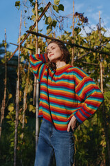 Fototapeta premium Woman in her mid 20s wearing a colorful striped sweater and jeans, smiling and leaning against a garden trellis among vines, relaxed lifestyle portrait in nature.