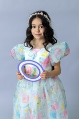 A cute girl in a floral party dress holding a purple oval frame. Studio portrait of a child with a festive headband on a light gray background