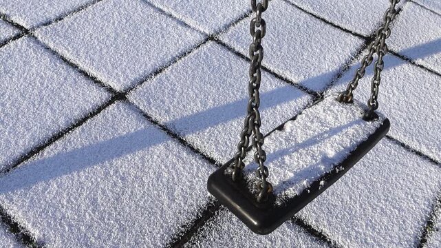 Top down slow motion video of a empty playground swing swinging and moving back and fourth during first snow day with white fluffy hale grainy snow around and on it. The swing has linked chain. Sunny.
