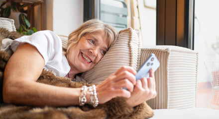 Senior woman smiling, lying on sofa, having a video chat on her smartphone at home