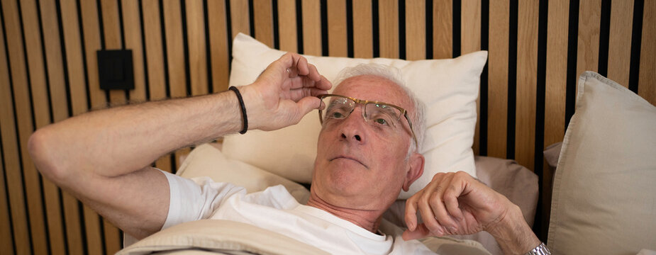 Senior man adjusting eyeglasses in bed, thinking about the day