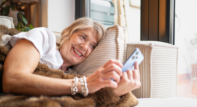 Senior woman smiling, lying on sofa, having a video chat on her smartphone at home - Powered by Adobe