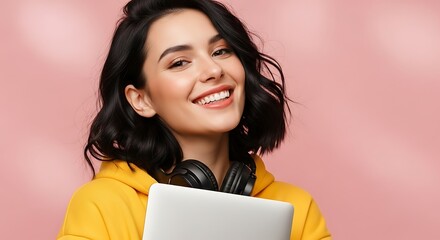 Cheerful student wearing a yellow hoodie hugging a laptop with headphones around her neck on a pink backdrop