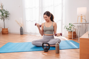 Woman workout at home using dumbbell and kettlebell on yoga mat, wearing activewear. Healthy lifestyle, strength training, wellness, fitness, and body toning in peaceful indoor space.