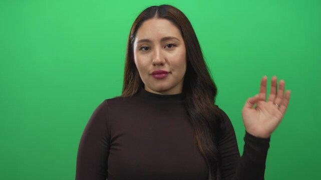 Hispanic young woman waves hand in studio with vibrant green backdrop while smiling gently; friendly greeting.