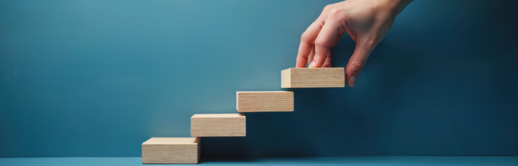Hand builds stairs with wooden blocks against blue background. This represents growth, progress, planning, and the start of a new project or career path.