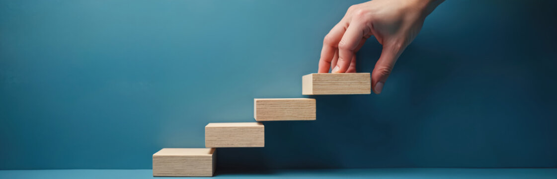 Hand builds stairs with wooden blocks against blue background. This represents growth, progress, planning, and the start of a new project or career path.