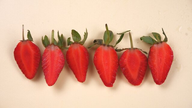 Six halved strawberries are arranged in a row, showcasing their vibrant red color and green stems against a neutral background. - Powered by Adobe