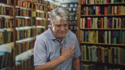 Man with hand on chest among bookshelves in a building, touching chest with fingers and slight smile; gratitude reflection.