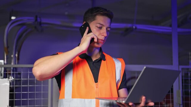 Dedicated professional in safety vest communicates on phone while operating laptop. Demonstrates focused problem-solving and efficient task management in industrial environment, showing commitment.