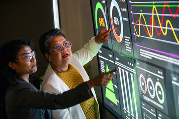 Two businesswomen in a modern office analyze interactive data visualizations on a large digital screen, focusing on analytics, performance monitoring, and strategic business efficiency.