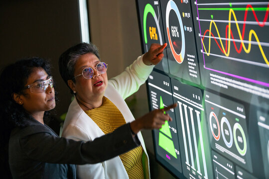 Two businesswomen in a modern office analyze interactive data visualizations on a large digital screen, focusing on analytics, performance monitoring, and strategic business efficiency.