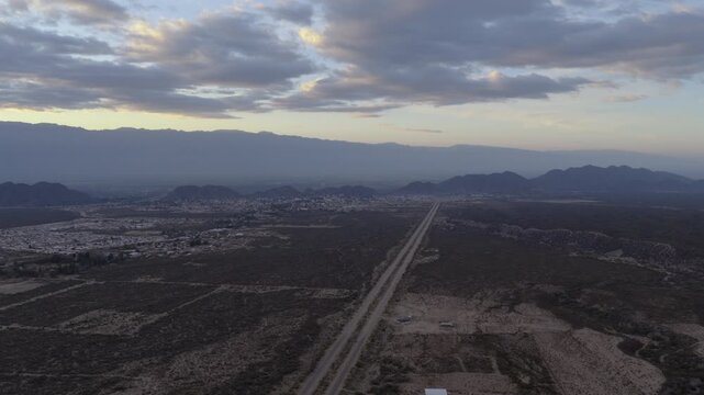 A drone flies forward above a long straight road at dawn, city and mountains fading into misty blue horizon.