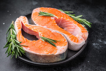 Raw salmon steaks with rosemary rest on a black plate against a dark textured background offering rich contrast for gourmet themes and recipe design.
