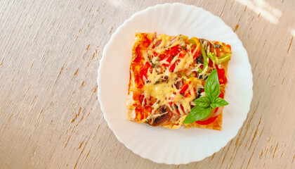 Slice of pizza with cheese, vegetables and basil on a white plate standing on a wooden table.