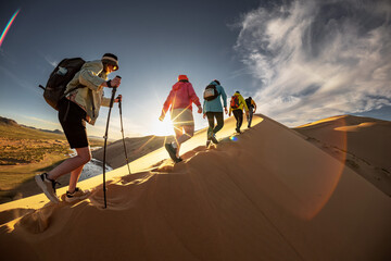 Group of young hikers goes uphill with backpacks and hiking poles to desert dune at sunset